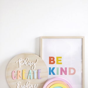 May include: A wooden round sign with the words "Play Create Explore" in colorful letters. A framed print with the words "Be Kind" in colorful letters. A rainbow-colored wooden arch and two wooden blocks with a tent and a deer.