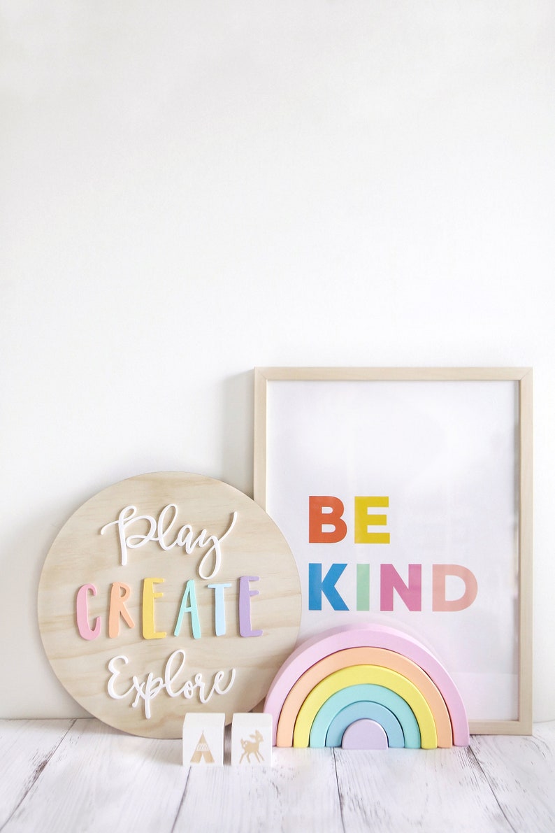 May include: A wooden round sign with the words "Play Create Explore" in colorful letters. A framed print with the words "Be Kind" in colorful letters. A rainbow-colored wooden arch and two wooden blocks with a tent and a deer.