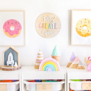 May include: A white shelf unit with three drawers, each labeled with a different word. The top drawer is labeled "Cars", the middle drawer is labeled "Blocks", and the bottom drawer is labeled "Arts & Crafts". The shelves are decorated with wooden toys, including a rocket, a rainbow, a mountain range, and a pink swan.