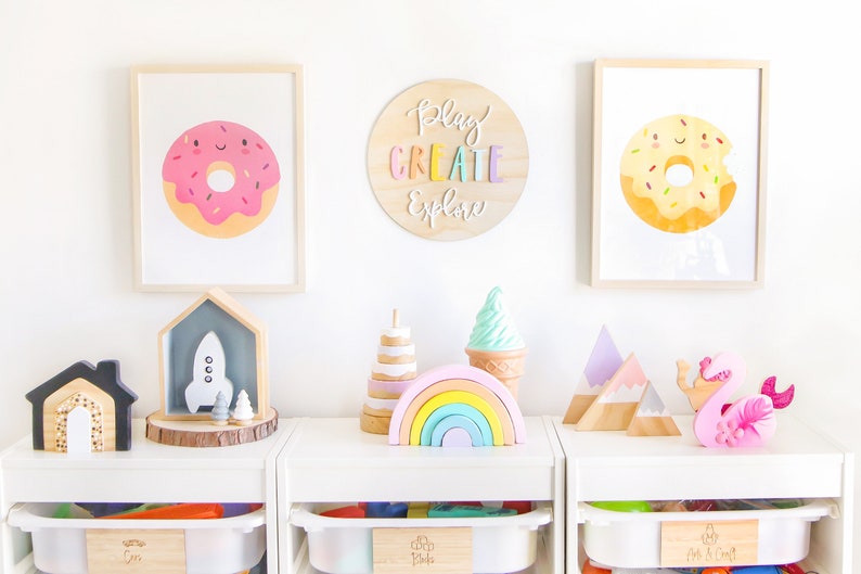 May include: A white shelf unit with three drawers, each labeled with a different word. The top drawer is labeled "Cars", the middle drawer is labeled "Blocks", and the bottom drawer is labeled "Arts & Crafts". The shelves are decorated with wooden toys, including a rocket, a rainbow, a mountain range, and a pink swan.