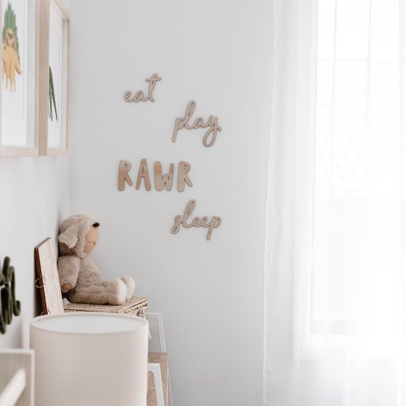 Op de afbeelding: Houten wanddecoratie met de woorden "eat", "play", "RAWR" en "sleep" in een witte kamer met een knuffeldier en een lamp.