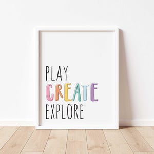 May include: White framed art print with the words "PLAY CREATE EXPLORE" in black and colorful letters. The artwork is displayed on a white wall above a wooden floor.