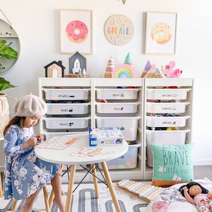 May include: A white bookshelf with clear plastic bins labeled with different themes, such as "Hot Wheels", "Blocks", "Art & Craft", "Trains", "Dolls", "Technology", "Puzzles", and "Airplanes". A young girl is playing with a game on a white table in front of the bookshelf. The game is a colorful set of cards with words on them. There is a wooden xylophone and a large, round, floral-patterned pillow on the floor. The wall behind the bookshelf is decorated with a wooden sign that says "Play Create Explore" and two framed pictures of donuts.