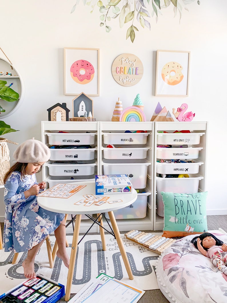 May include: A white bookshelf with clear plastic bins labeled with different themes, such as "Hot Wheels", "Blocks", "Art & Craft", "Trains", "Dolls", "Technology", "Puzzles", and "Airplanes". A young girl is playing with a game on a white table in front of the bookshelf. The game is a colorful set of cards with words on them. There is a wooden xylophone and a large, round, floral-patterned pillow on the floor. The wall behind the bookshelf is decorated with a wooden sign that says "Play Create Explore" and two framed pictures of donuts.