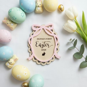 May include: A wooden Easter sign with the text "Olivia's First Easter 2026" in a decorative frame, surrounded by pastel-coloured Easter eggs, bunny figurines, and white tulips. A golden egg is also present.