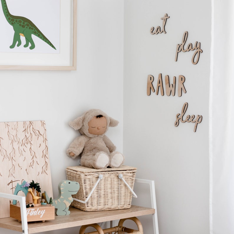 Op de afbeelding: Een wit rek met een rieten mand, een houten speelgoed dinosaurus, een houten muziekdoos en een knuffeldier dat een lammenkostuum draagt. De muur achter het rek heeft houten letters die "eat play RAW sleep" spellen.