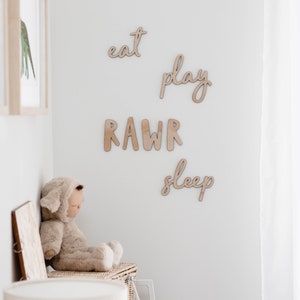 Op de afbeelding: Houten wanddecoratie met de woorden "eat", "play", "rawr" en "sleep" in een cursief schrift. Een knuffeldier zit op een plank onder de decoratie.