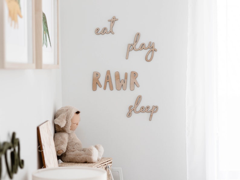 Op de afbeelding: Houten wanddecoratie met de woorden "eat", "play", "rawr" en "sleep" in een cursief schrift. Een knuffeldier zit op een plank onder de decoratie.