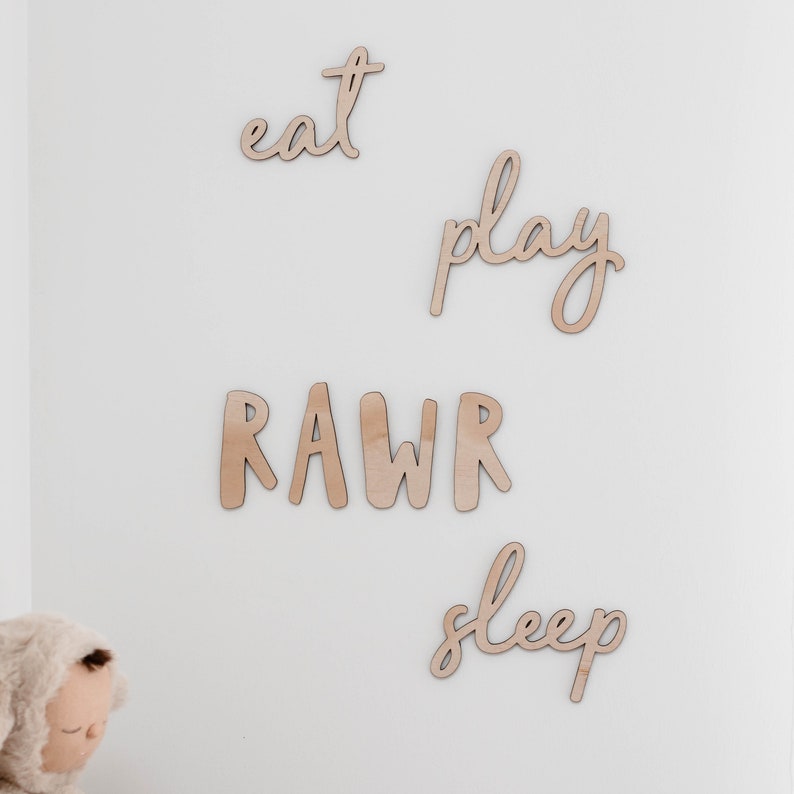 Op de afbeelding: Houten wanddecoratie met de woorden "eat", "play", "rawr" en "sleep" geschreven in een cursief schrift.
