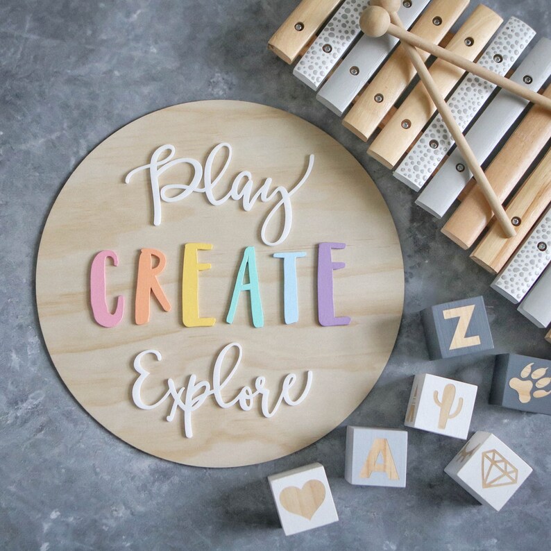 May include: A wooden round sign with the words "Play Create Explore" in colorful letters. The sign is surrounded by wooden blocks with letters and symbols, and a wooden xylophone.