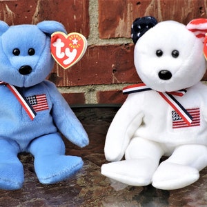 May include: Two plush beanie baby bears, one blue and one white, both wearing red, white, and blue ribbon and a small American flag patch. The bears are sitting on a brown surface.