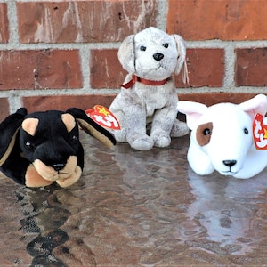 Könnte beinhalten: Drei Plüschhunde, ein schwarz-brauner Dackel, ein weißer Bullterrier und ein grau-weißer Hund, alle mit roten Ty-Etiketten.