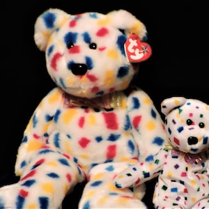 May include: Two white plush teddy bears with colourful confetti-like patterns in red, blue, and yellow. The larger bear has a bow tie and a red heart-shaped tag. The smaller bear also has a bow tie and a tag.