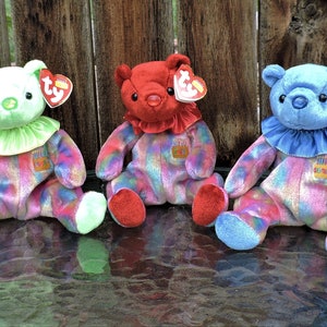 May include: Three Ty Beanie Babies in a rainbow swirl pattern. The bears are green, red, and blue with a birthday cake patch on their chests. Each bear has a Ty tag attached to its ear.