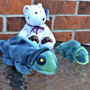 Könnte beinhalten: Drei Plüschtiere: ein weißer Bär mit bunter Stickerei, ein blaues und grünes Chamäleon und ein grüner Frosch. Die Spielzeuge sitzen auf einem Glastisch.