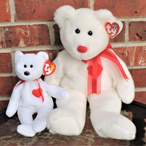 May include: Two white plush teddy bears with red hearts on their chests. The larger bear has a red ribbon around its neck. Both bears have Ty tags attached to their ears.