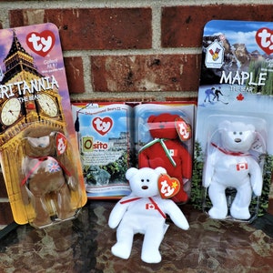 Op de afbeelding: Drie Ty Beanie Baby pluche speelgoed in hun originele verpakking. De speelgoed zijn Britannia de Beer, Osito de Beer en Maple de Beer. De verpakking toont afbeeldingen van de Big Ben klokkentoren, het Colosseum en een Canadees berggebied.