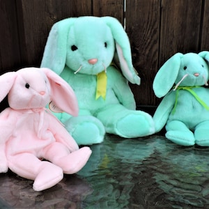 May include: Three plush bunny toys. One is pink, one is light blue, and one is a darker shade of blue. All three have long ears and are sitting on a textured surface.