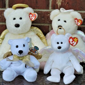 May include: Four white plush angel teddy bears with golden and iridescent wings. The bears have black eyes and noses and are wearing haloes. The bears are sitting on a table in front of a brick wall.