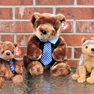 Puede incluir: Tres juguetes de peluche: un oso marrón con una camisa azul y una corbata de lunares azules y blancos, un oso marrón más pequeño con una camisa azul y una corbata de lunares azules y blancos, y un perro marrón y gris con un sombrero gris.