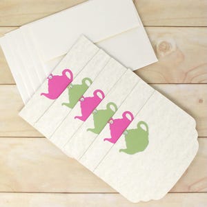 May include: Cream-colored stationery set featuring a scalloped edge design. Each card has a pink or green teapot graphic with a small gem. Includes matching cream-colored envelopes. Perfect for sending notes or invitations.