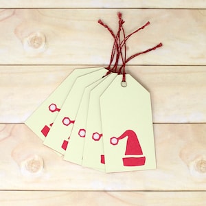 May include: Five cream-coloured gift tags with red string attached. Each tag has a red Santa hat design with white trim. The tags are arranged in a fan shape.