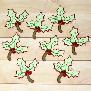 May include: Eight decorative holly leaf cutouts. Each leaf is light green with a red outline and stem, and red berries. The leaves are arranged on a light brown wooden surface.