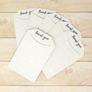 May include: A group of off-white paper envelopes, each with a curved top flap. The phrase "thank you" is printed in a cursive font across the top of each envelope. The envelopes are arranged on a light-coloured wooden surface.