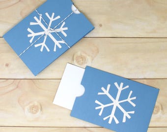May include: Two light blue gift card holders with white snowflake designs. One holder is closed with a white string, and the other is open, revealing a white card. The holders are on a light brown wooden surface.