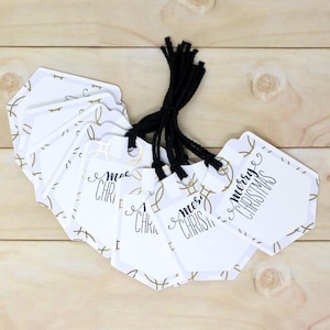 May include: A collection of white Christmas gift tags with gold geometric accents and black string. Each tag has the words "merry CHRISTMAS" in black script. The tags are arranged on a light wood surface.