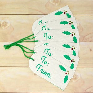 May include: A collection of white gift tags with green text reading "To:" and "From:". Each tag features a green holly leaf design with red and gold accents. The tags are bound together with green string.