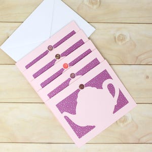 May include: Pink greeting cards with a teapot silhouette, purple polka dot accents, and decorative buttons. A white envelope rests on a light wood surface. The cards are arranged in a fan shape.