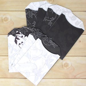 May include: A collection of decorative paper envelopes. The envelopes feature a black and white floral design with a leaf pattern. The envelopes are arranged on a light brown wooden surface.