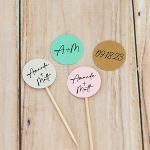 May include: Four round toppers on wooden sticks, perfect for a wedding or event. The toppers are in various colors: mint green, kraft brown, light pink, and off-white. Each topper has black script text, including initials, names, and a date.