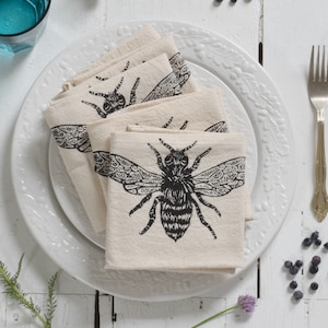 May include: Four white linen napkins with a black bee design are folded and stacked on a white plate. The plate is on a white wooden table with blue berries scattered around it.