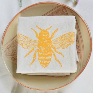 May include: A folded white linen tea towel with a yellow bee design printed on it. The towel is sitting in a light brown ceramic bowl.