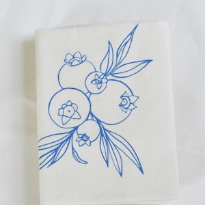 May include: A folded, off-white cloth napkin featuring a blue line drawing of blueberries and leaves. The illustration depicts several blueberries clustered together with delicate stems and foliage, creating a simple, elegant design.