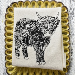 May include: A black and white illustration of a Highland cow printed on a white tea towel. The tea towel is folded and placed on a yellow ceramic tray with a scalloped edge.
