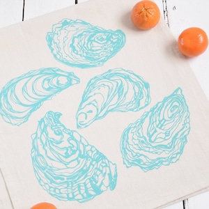 May include: A white kitchen towel with a blue oyster design. The towel is decorated with five blue oyster shells. There are three orange clementines on the white wooden surface.