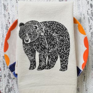 May include: A white kitchen towel with a black and white illustration of a bear. The towel is folded and placed on top of a colorful ceramic plate.