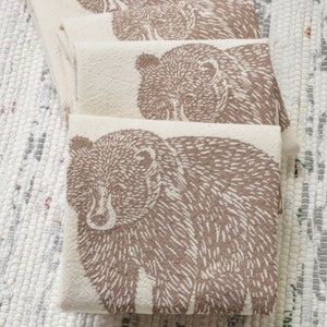 May include: Set of four brown and white kitchen towels with a bear print design. The towels are folded and stacked on top of each other.