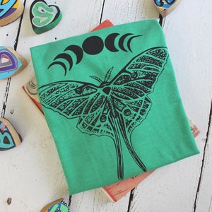 May include: A green t-shirt with a black and white illustration of a moth and a crescent moon design. The shirt is folded and lying on a white wooden surface.