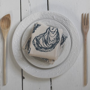 May include: A white ceramic plate with a raised design, featuring a folded linen napkin with a blue oyster print. The plate is set on a white wooden surface with a wooden spoon and fork.
