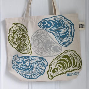 May include: Cream-colored tote bag with a repeating oyster shell design in shades of blue, green, and gray. The bag has long, beige handles and the text "HEARTHE HARROW" is printed on the bottom right corner.