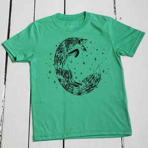 Puede incluir: Una camiseta verde con un gr&aacute;fico negro de un zorro acurrucado en forma de media luna. El zorro est&aacute; rodeado de peque&ntilde;as estrellas.