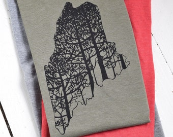 May include: Three t-shirts stacked on top of each other. The top shirt is olive green with a black silhouette of trees in the shape of the state of Maine. The middle shirt is red. The bottom shirt is gray.