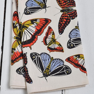 May include: A folded cream-colored kitchen towel featuring a repeating pattern of colorful butterflies. The butterflies have black, red, yellow, and blue wings. The towel is made of a soft, absorbent fabric, perfect for drying dishes or wiping surfaces.
