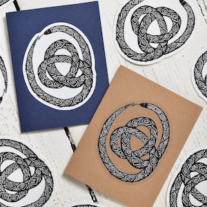 May include: Cards and stickers with a black and white snake design. The snake is coiled in a circular pattern with a geometric diamond pattern. The cards are navy blue and tan, and the stickers are white.