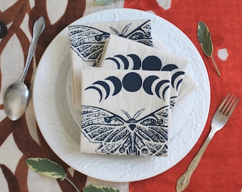 May include: A set of three white cotton napkins with blue and black printed designs. The designs include a moth, a crescent moon, and a full moon. The napkins are folded and stacked on a white plate.