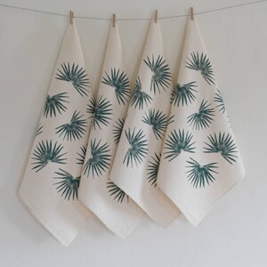 May include: Four white kitchen towels with a green palm leaf print hanging on a clothesline.